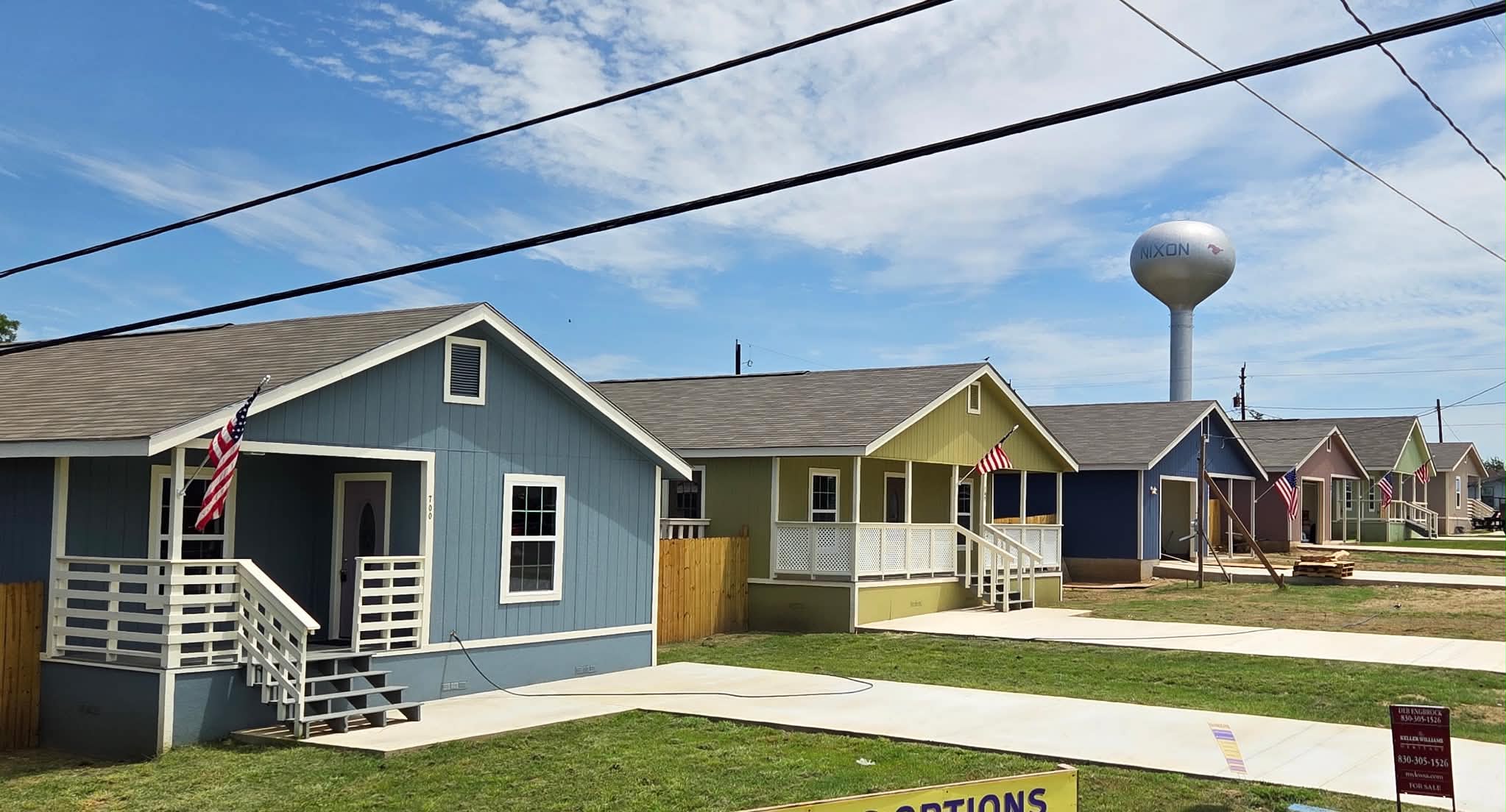 A row of newly built single-storey homes in blue, olive, navy and pink with white porch rails and American flags, the Nixon water tower behind