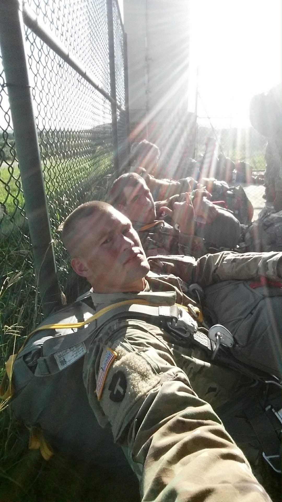 Paratroopers in jump harness lined up along a chain-link fence in strong sun, from Aurey's Army service