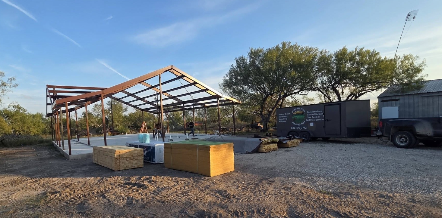 Dawn Till Dusk Construction work trailer parked beside a new steel frame on a fresh concrete slab, framing lumber stacked on site