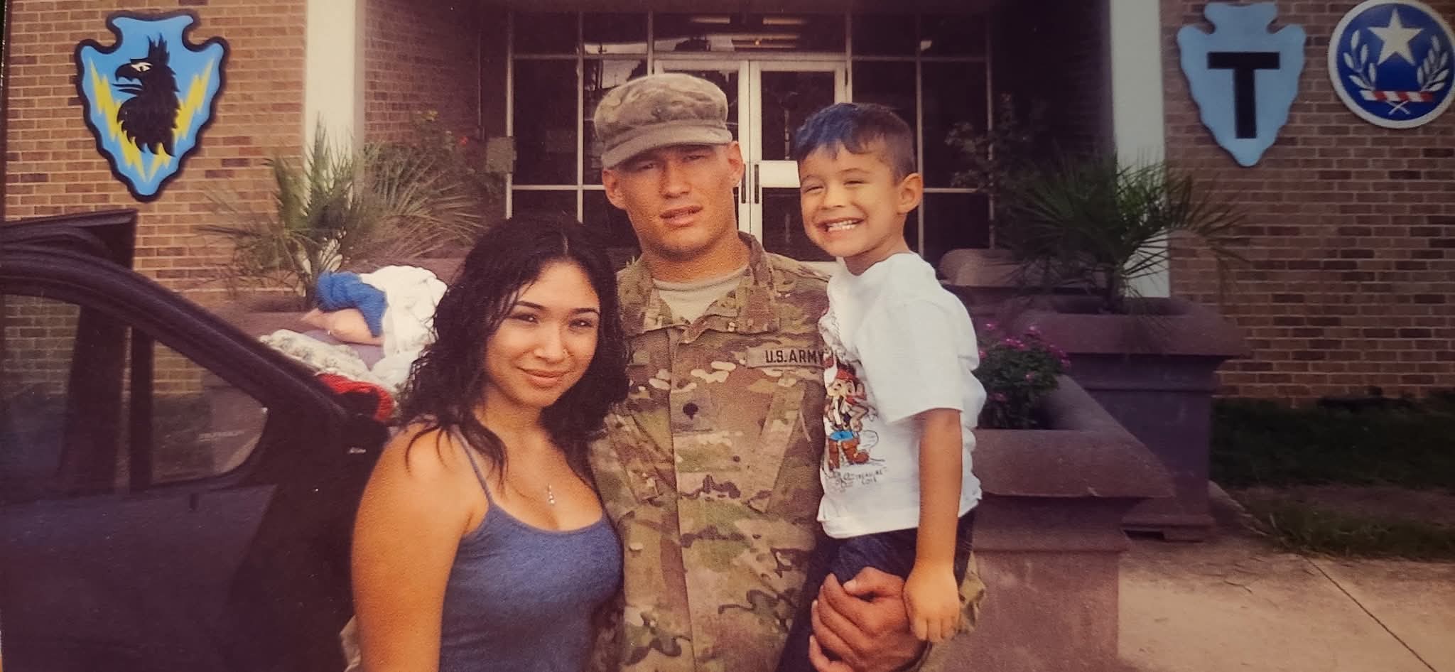 Aurey Kunschik in Army camouflage uniform with his family outside a brick building hung with unit crests