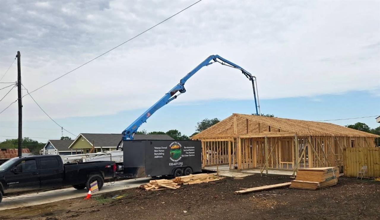 Concrete pump boom reaching over a newly framed house, with the Dawn Till Dusk Construction trailer alongside